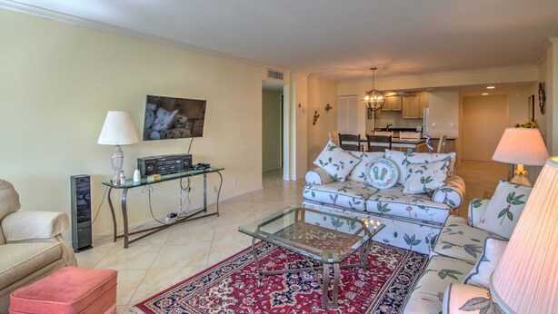 A living room with a sofa, a glass coffee table, a red patterned rug, a television, and a kitchen in the background. - Charming Coastal Living: Gulf Views Await (Naples)