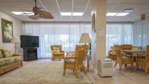 A living room with a flat-screen TV, wicker furniture, a ceiling fan, and a large window with curtains. - Charming Coastal Living: Gulf Views Await (Naples)