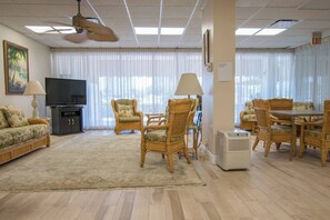 Ein Wohnzimmer mit Flachbildfernseher, Rattanmöbeln, Deckenventilator und einem großen Fenster mit Vorhängen.
