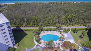 A resort with a central swimming pool, surrounded by lush greenery and a view of the ocean. - Charming Coastal Living: Gulf Views Await (Naples)