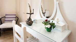 A bathroom with a white vanity, a mirror, a chair, and a small table with a vase of flowers. - Lydia Apartment (Corfu)