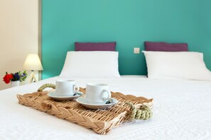 A neatly made bed with two white cups and saucers on a woven tray, a teal wall, and a white lamp with flowers.