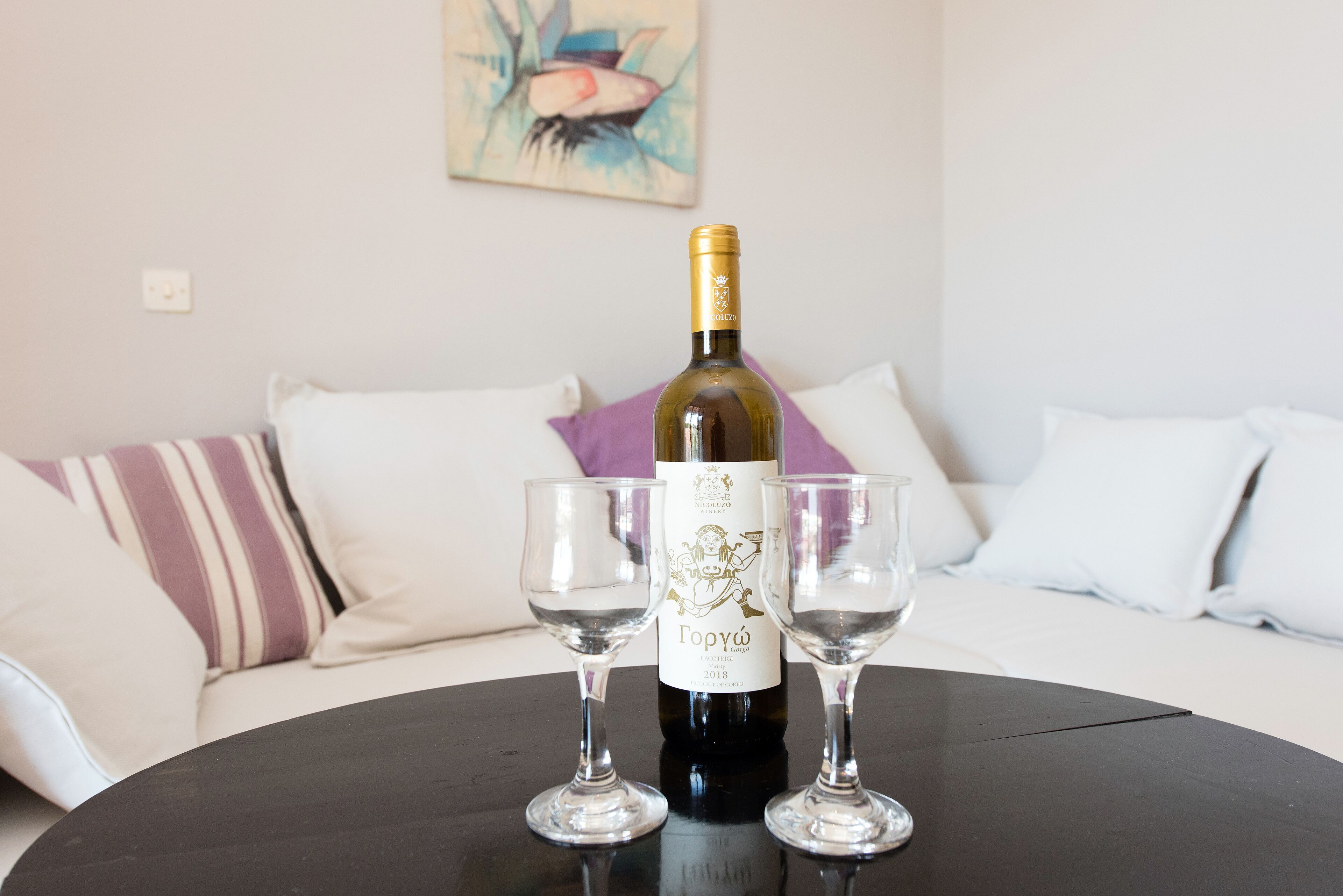 A bottle of wine on a table with two glasses, a sofa with striped pillows, and a painting on the wall.