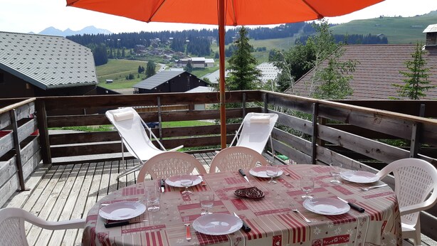 A wooden deck with a table set for four, surrounded by white chairs, overlooking a scenic view of houses and mountains.