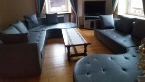 Un soggiorno con un divano componibile, un poggiapiedi rotondo, un tavolino da caffè, una TV a schermo piatto e una lampada a sospensione.