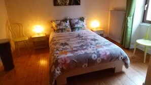 Una camera da letto con pavimento in legno, un letto con biancheria a fiori, due lampade da comodino, una poltrona verde e un quadro appeso al muro.