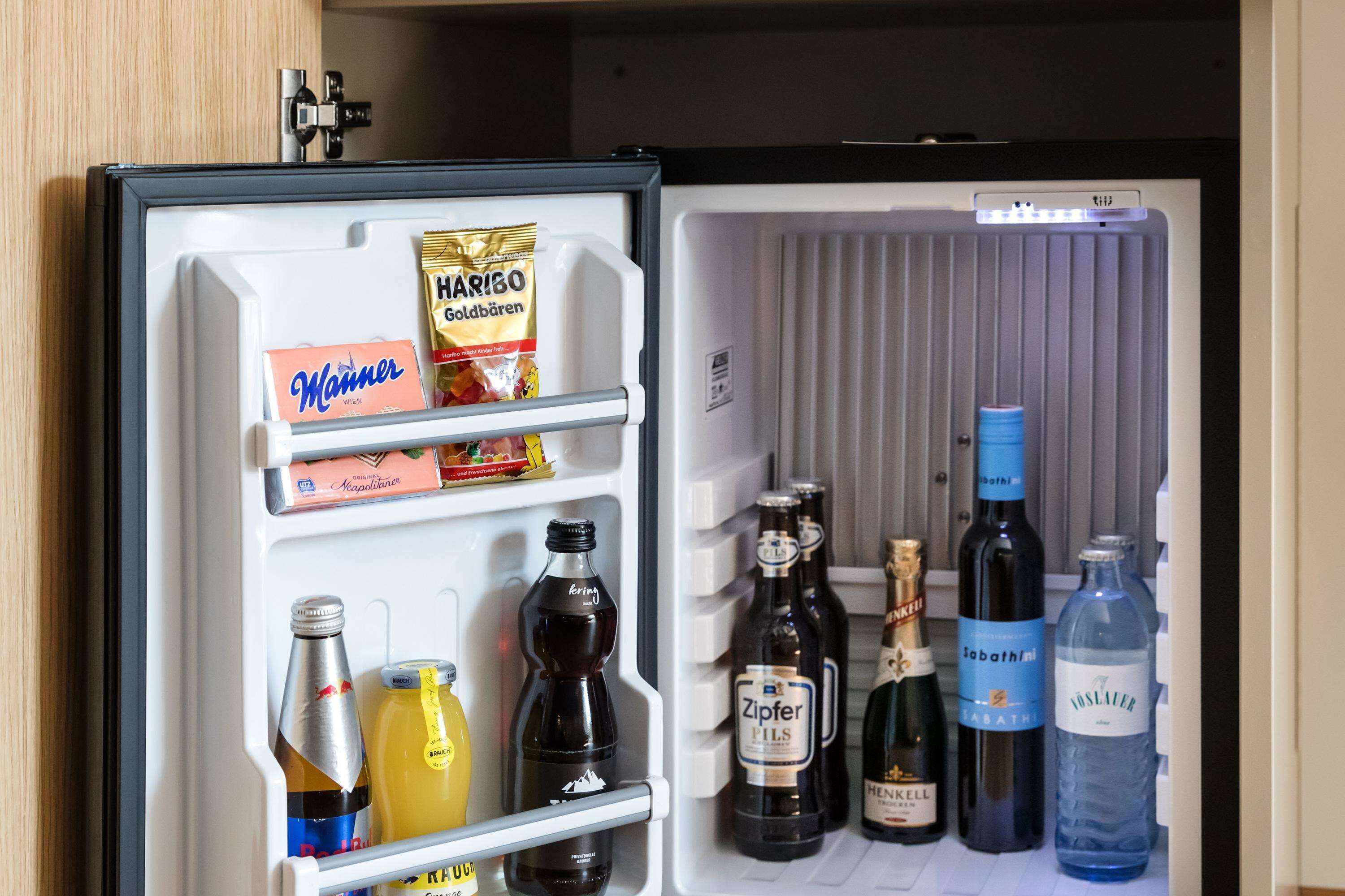 Un réfrigérateur ouvert contenant divers aliments et boissons, notamment une boîte de chocolats Hammer, un paquet de bonbons Haribo Goldbären et plusieurs bouteilles de bière et d’eau.