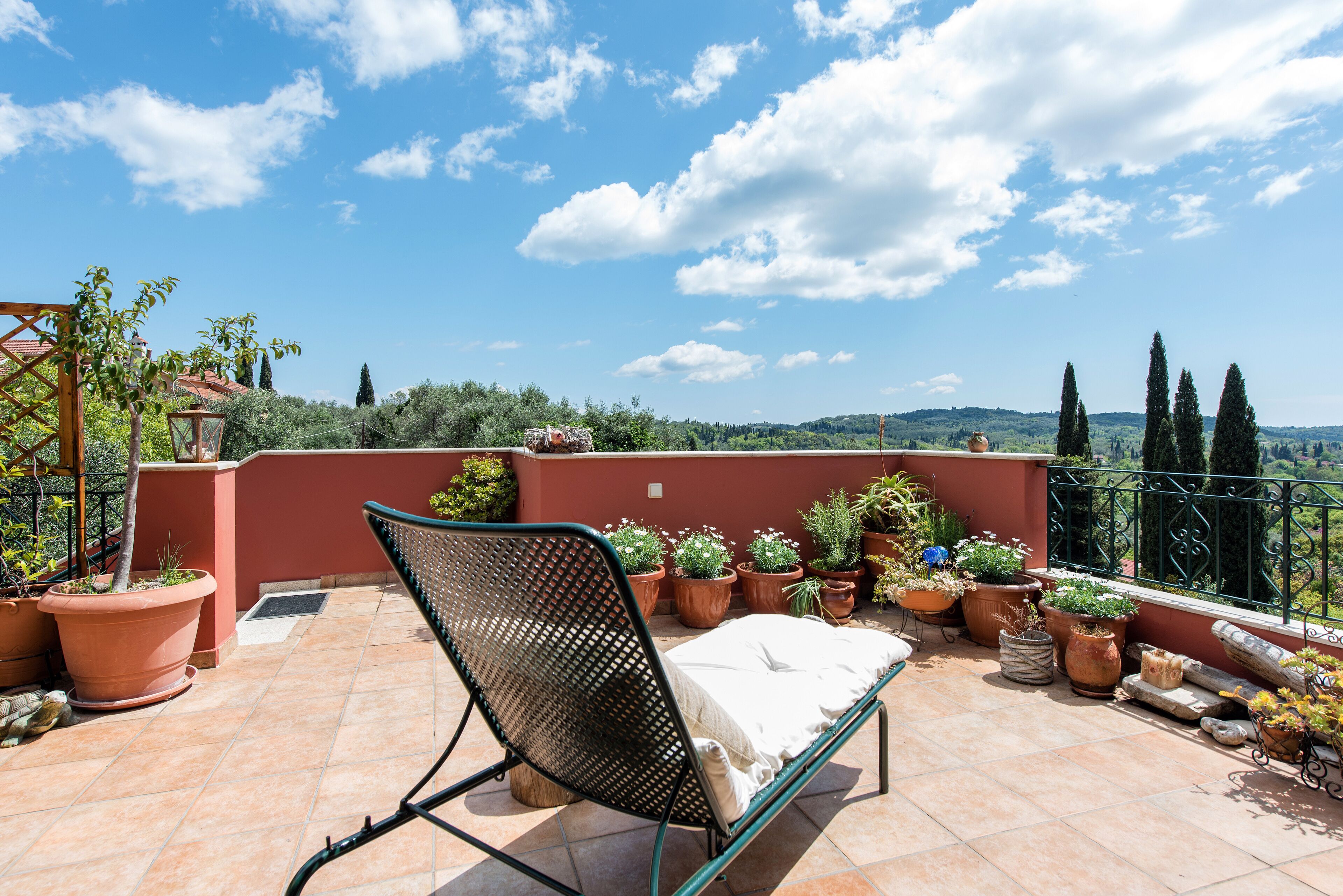 Una terraza con una silla de metal y una cama pequeña, rodeada de plantas en macetas y una vista panorámica de colinas.