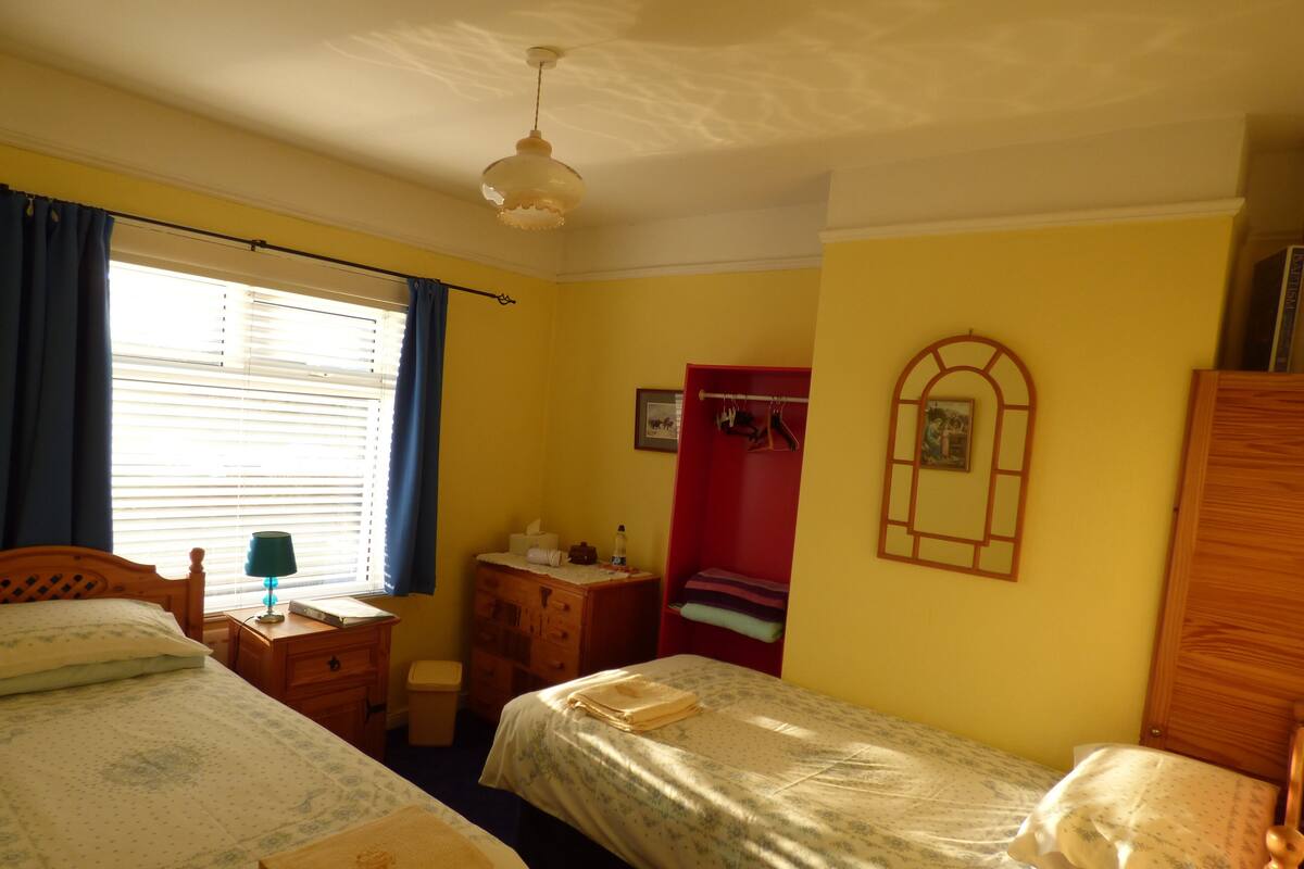 A room with two beds, a wooden wardrobe, a small desk, a mirror, and a window with curtains.