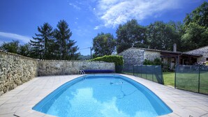 Una piscina rettangolare con un muro in pietra su un lato e, sullo sfondo, un’area recintata con prato e alberi.