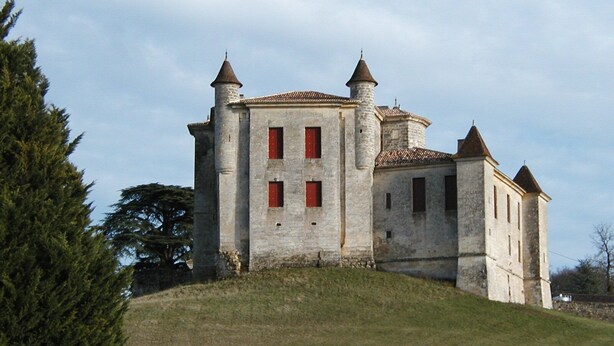 Un edificio storico in pietra con diverse torri e finestre rosse, immerso in una collinetta erbosa.