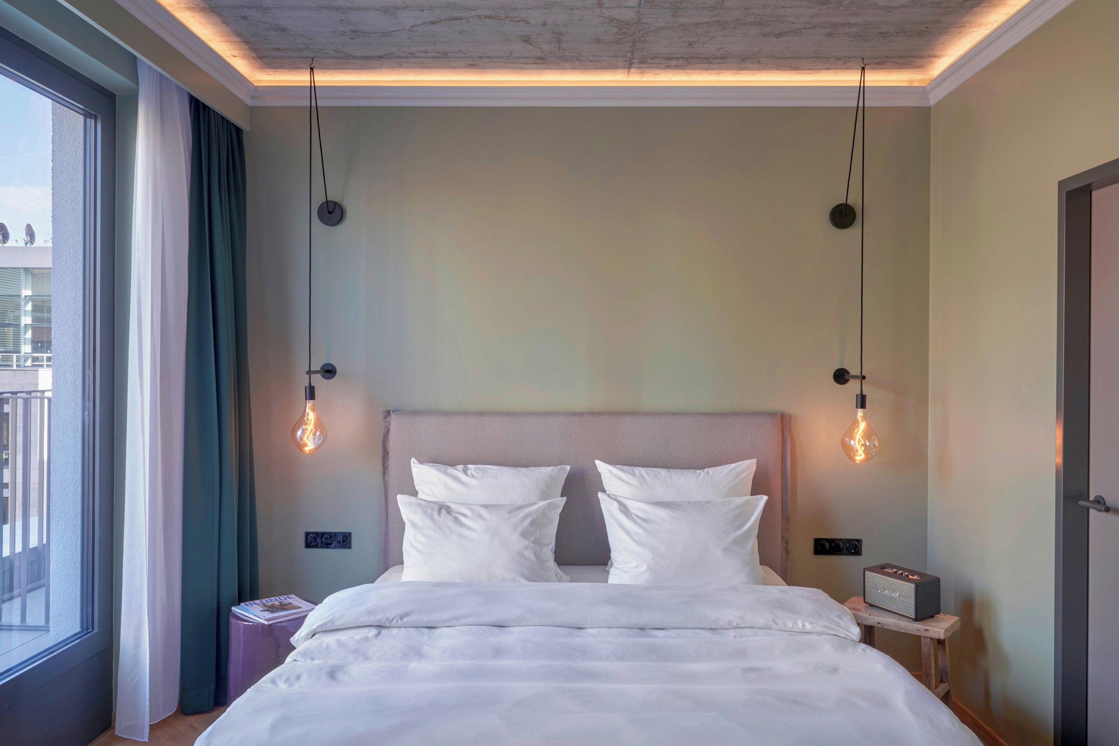 A modern hotel room with a large bed, two bedside tables, a nightstand, a window with curtains, and a ceiling with exposed beams.