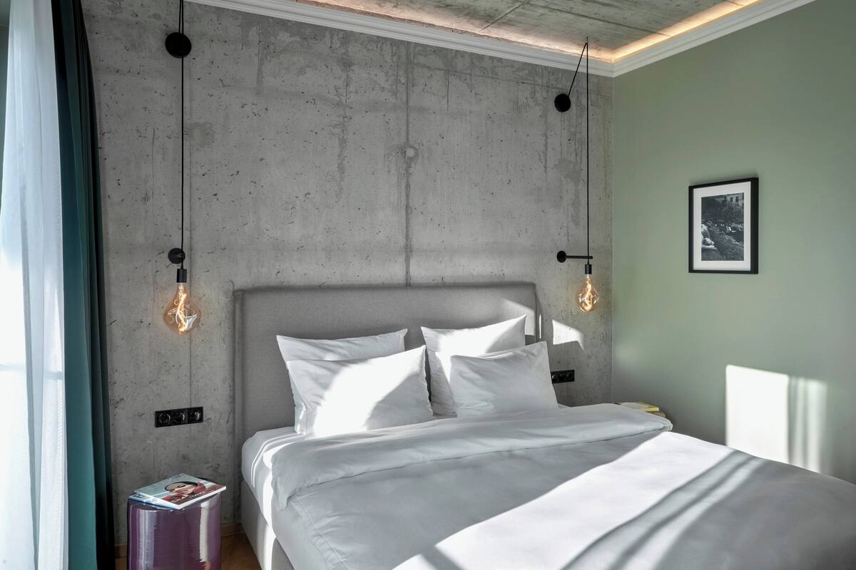 A modern hotel room with a concrete accent wall, a large bed with white linens, and pendant lights.