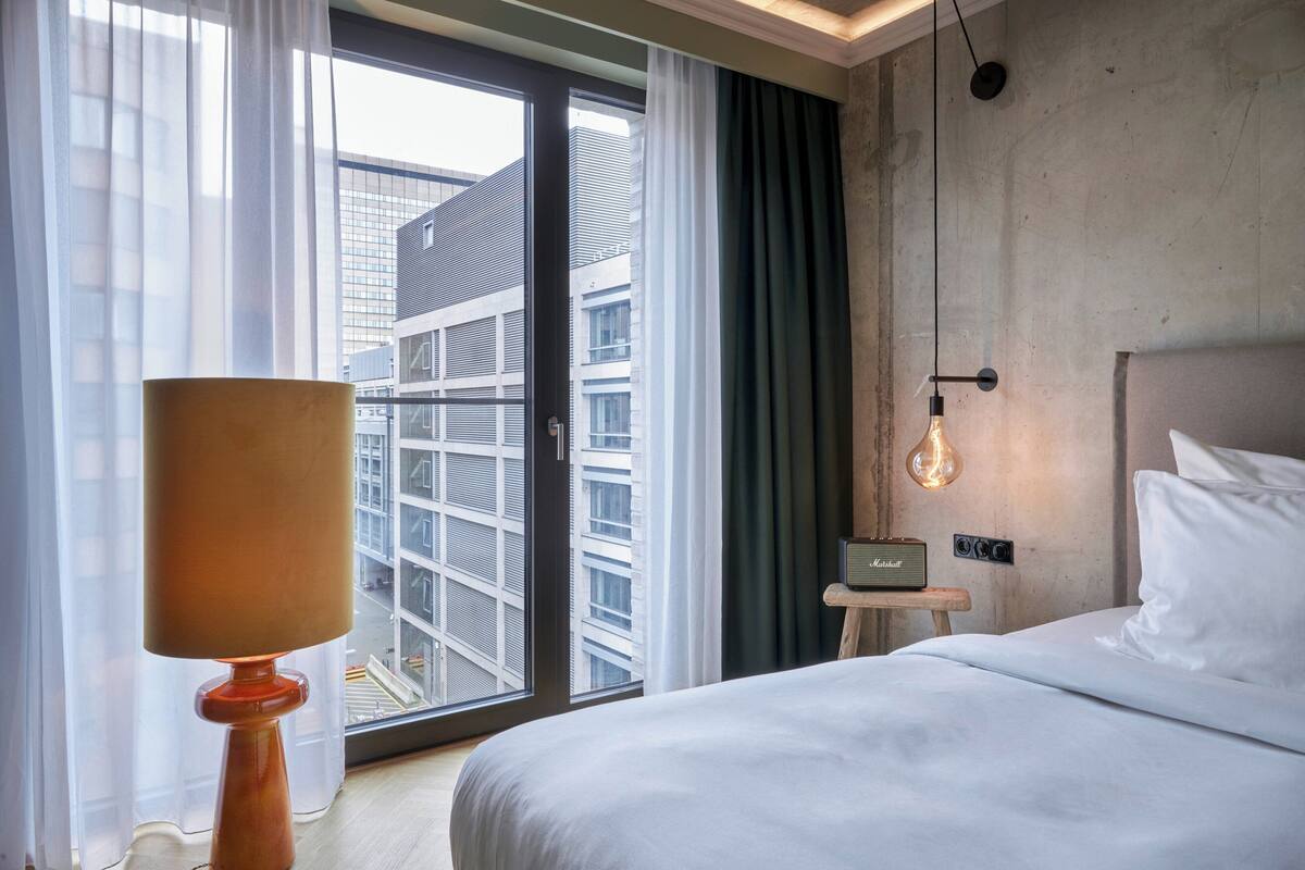 A hotel room with a bed, a lamp, a radio, and a view of a building through the window.