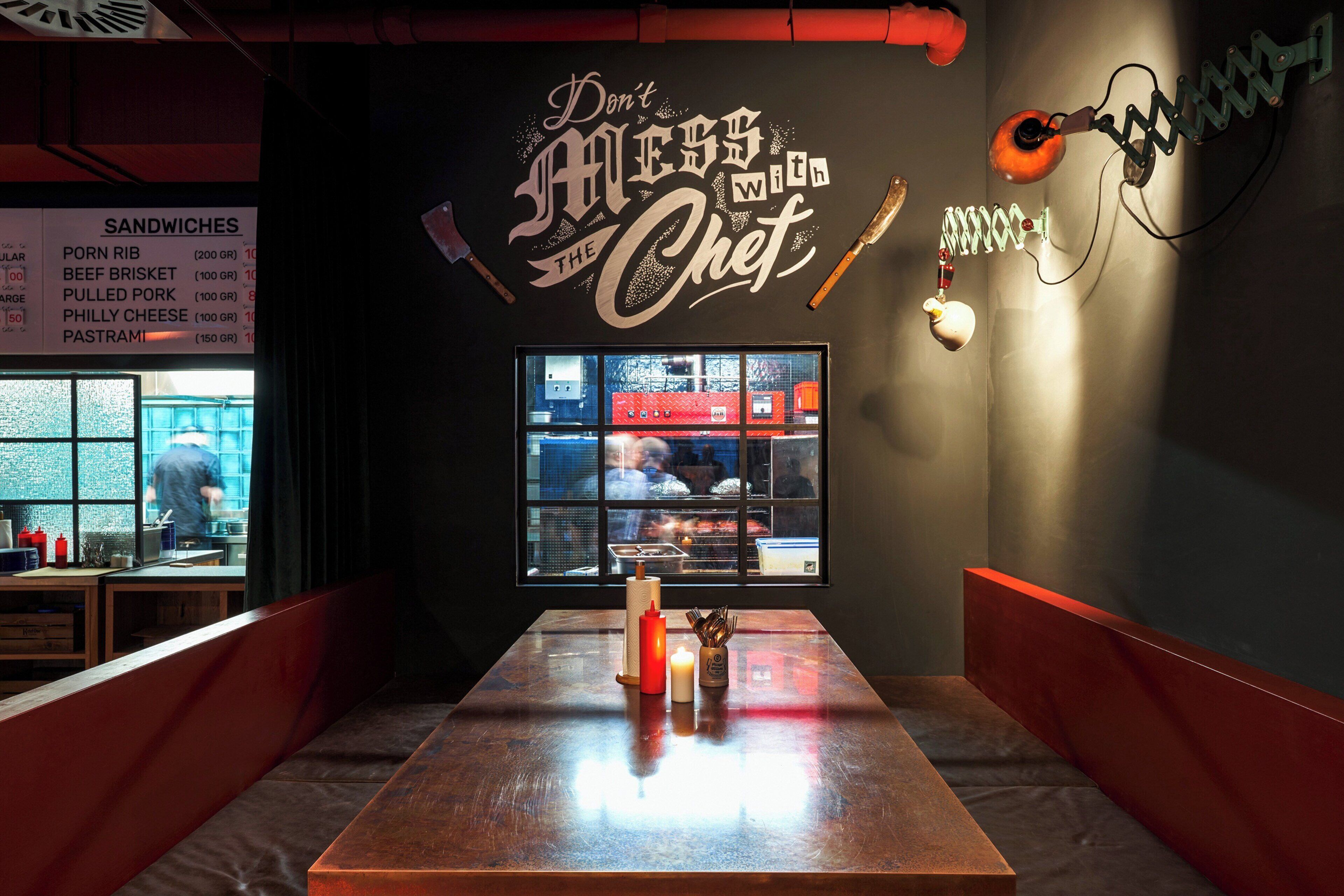 A restaurant interior with a menu board on the left, a wall with a 'Don't Mess with the Chef' sign, and a window view of the kitchen.