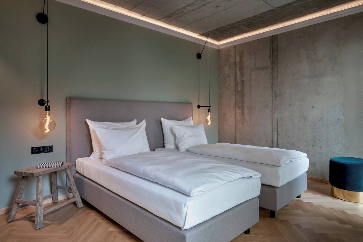 A modern bedroom with two beds, a wooden stool, and a round ottoman.