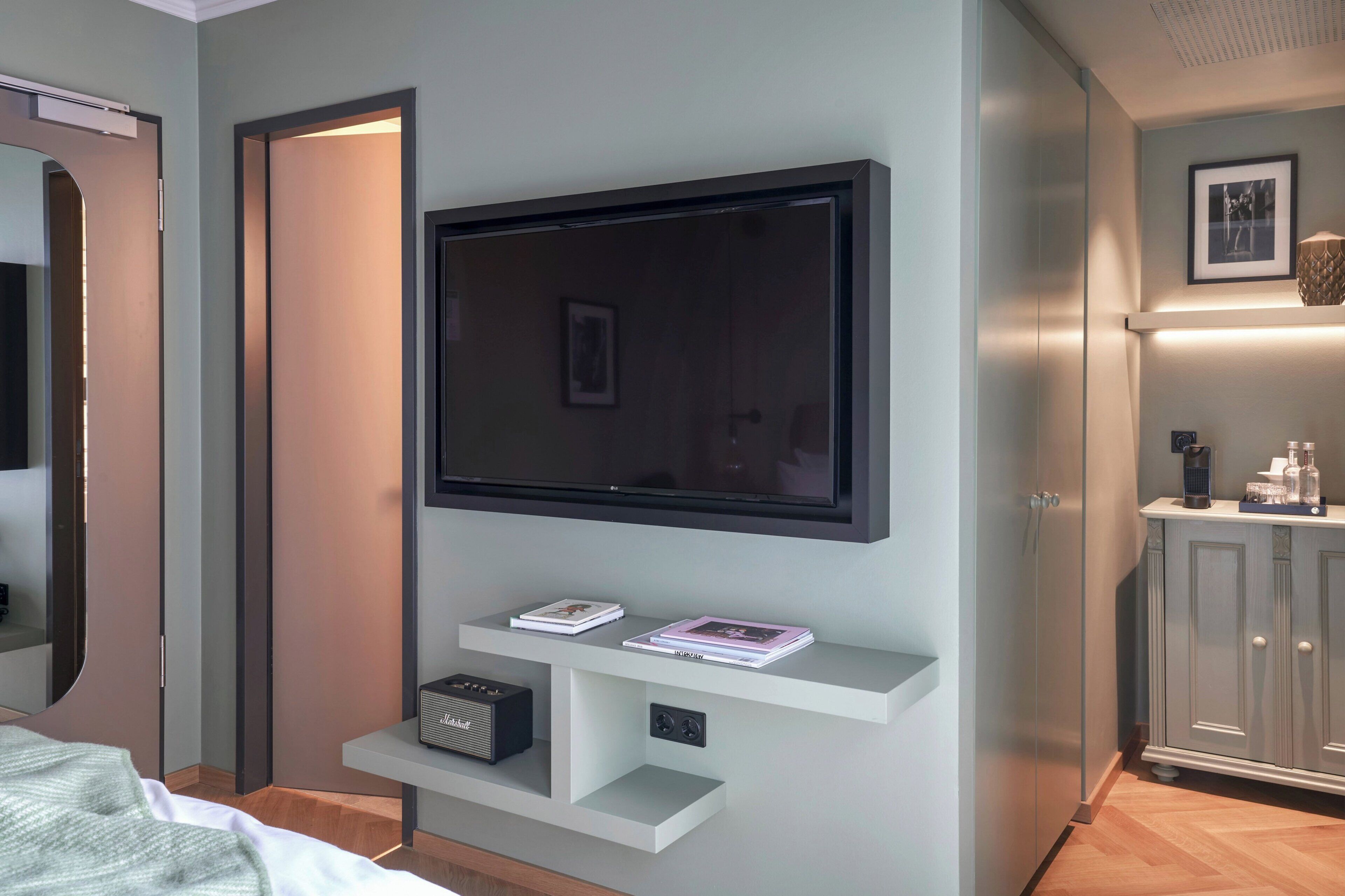 A modern hotel room with a flat-screen TV mounted on the wall, a small shelf with books and a radio, and a cabinet with toiletries.