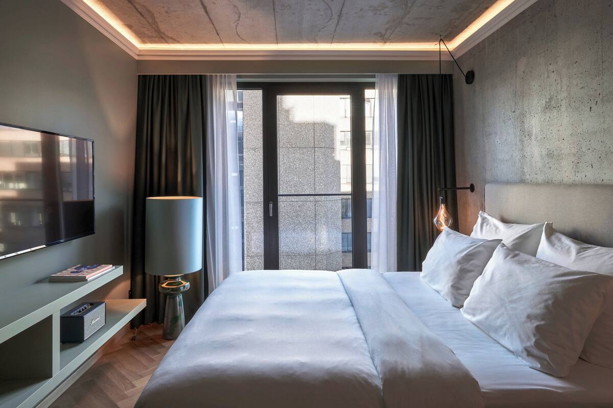 A modern hotel room with a large bed, a flat-screen TV, a bedside table with a lamp, and a view of the city through a glass door.