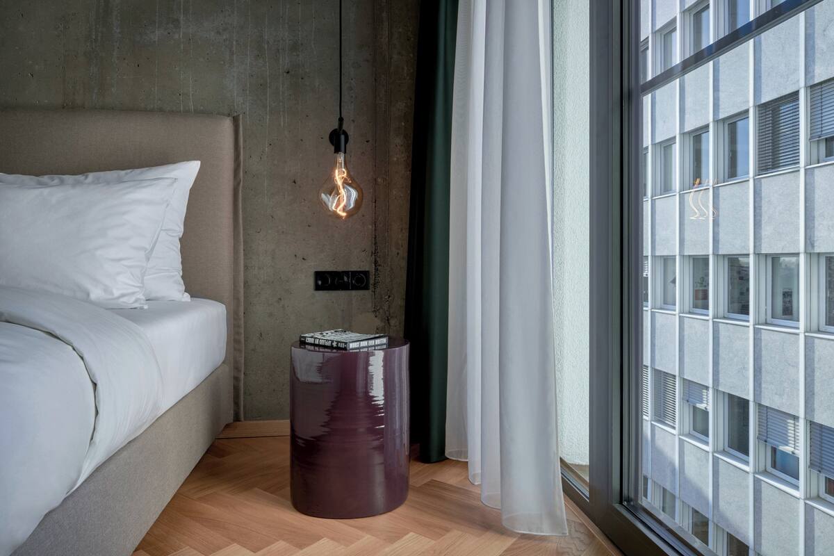 A bedroom with a bed, a light bulb hanging from the ceiling, a trash bin, and a window with blinds.