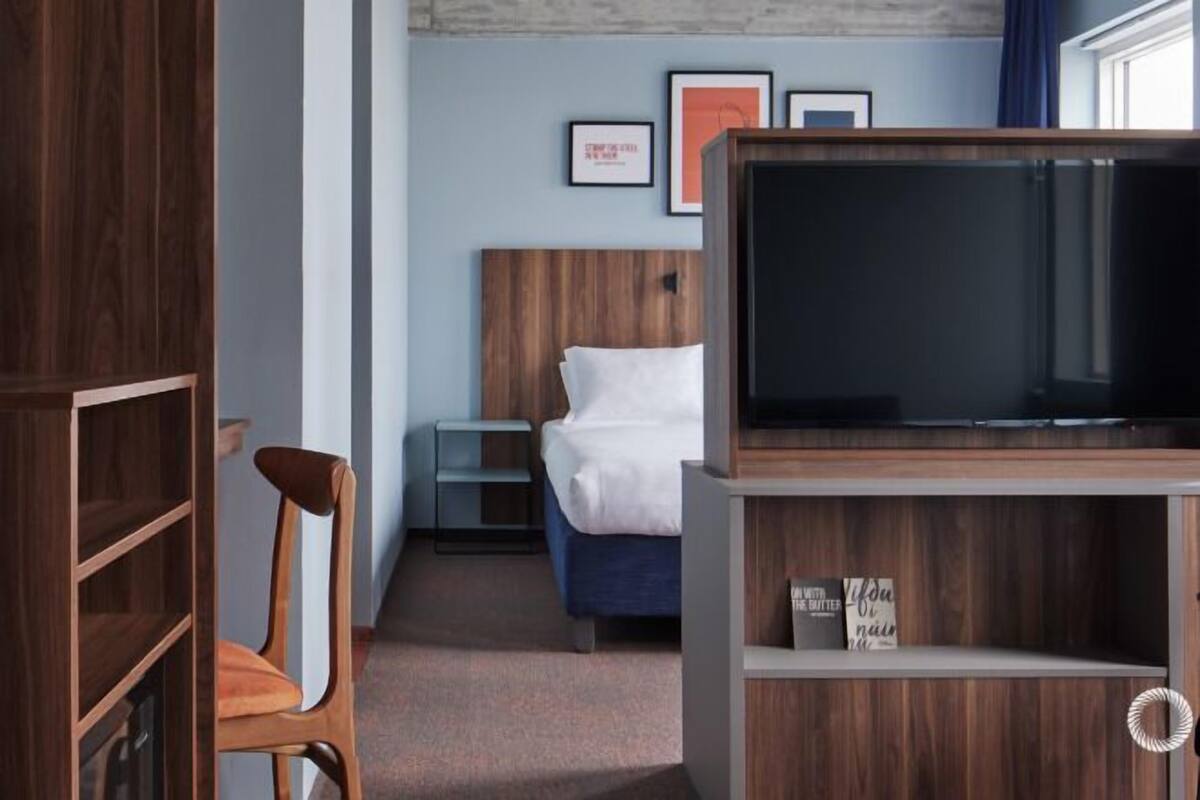 Ein Hotelzimmer mit einem Bett, einem Fernseher, einem Holzstuhl, einem Nachttisch und gerahmten Bildern an der Wand.