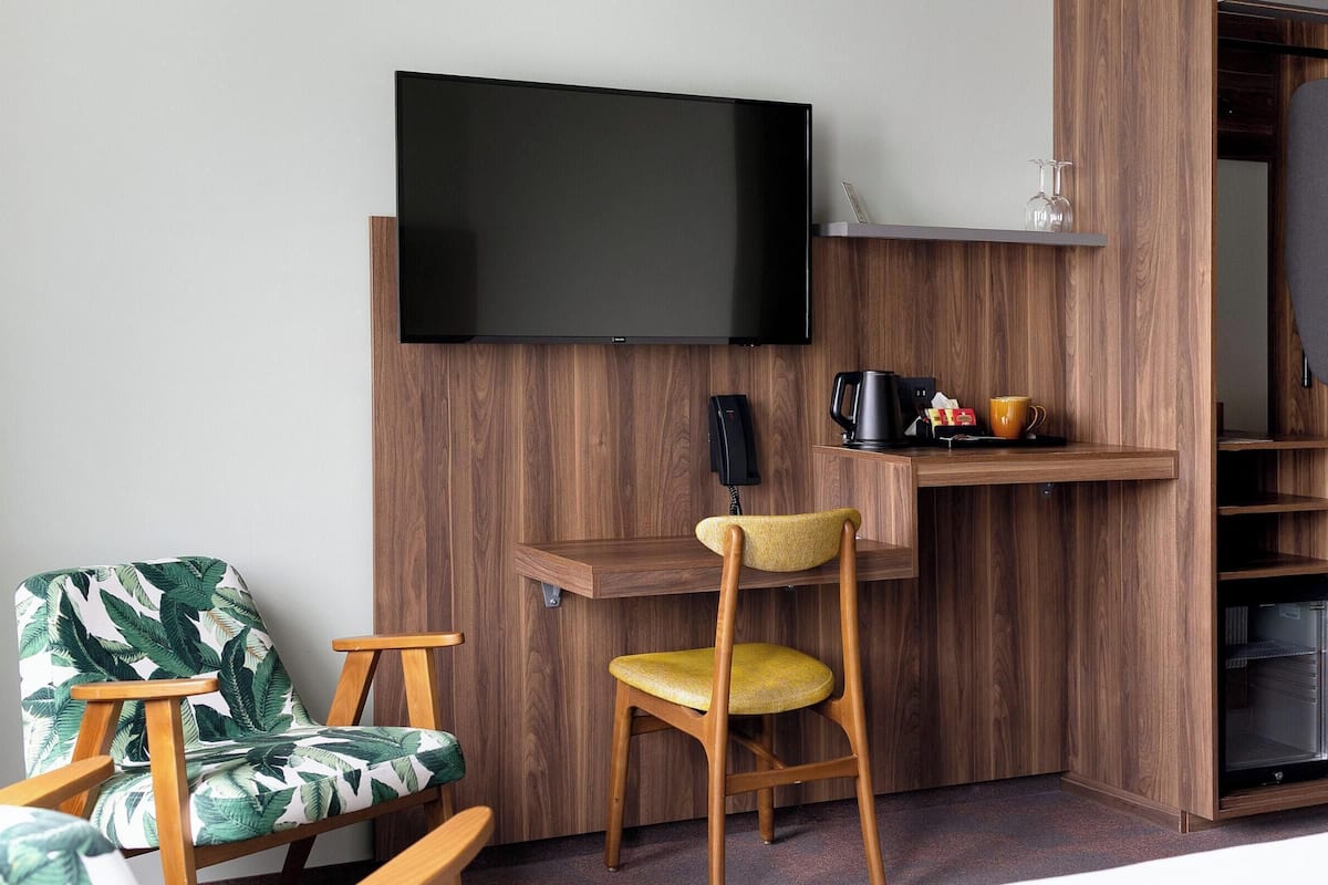 Ein gemütlicher Raum mit Holzwand, einem Flachbildfernseher über einem Holztisch, einem Sessel mit Blattmuster-Kissen und einer kleinen Ablage mit Kaffeemaschine und Tasse.