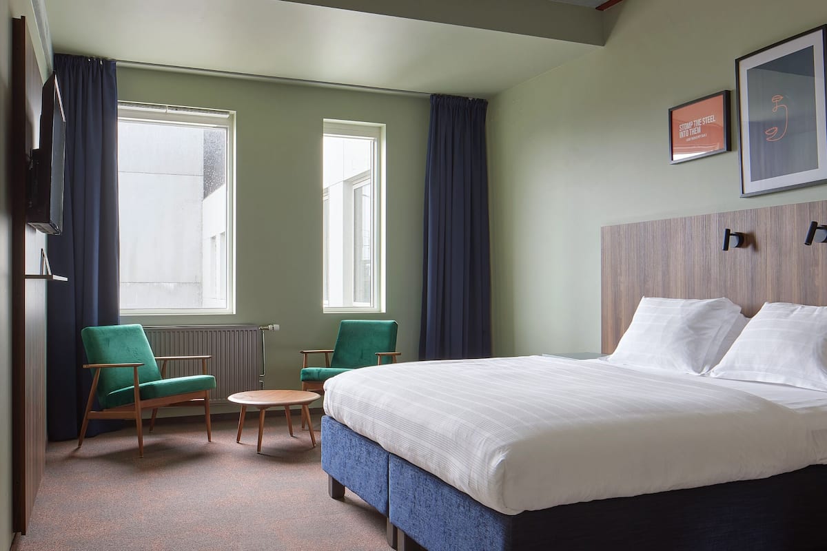 Ein Hotelzimmer mit einem großen Bett, zwei Sesseln, einem kleinen Tisch und zwei mit Vorhängen versehenen Fenstern.
