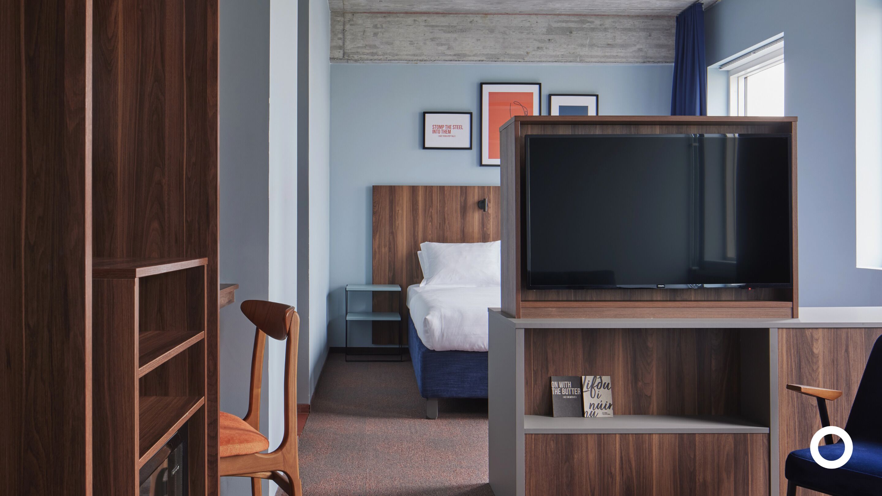 A hotel room with a wooden wardrobe, a bed with white bedding, a television on a wooden stand, and a small shelf with books.
