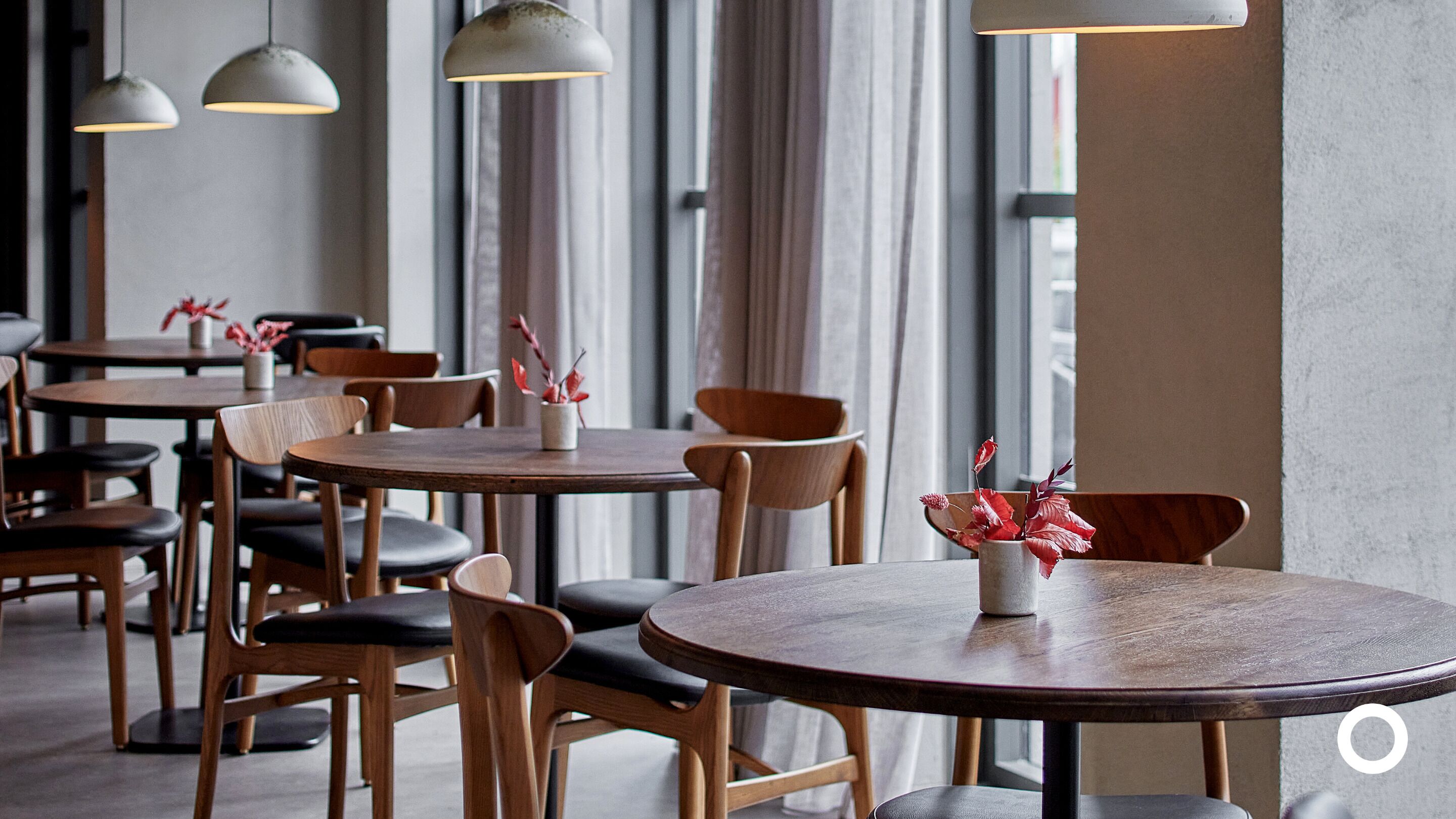A modern dining area with round tables and wooden chairs, featuring pendant lighting and a vase with red flowers.