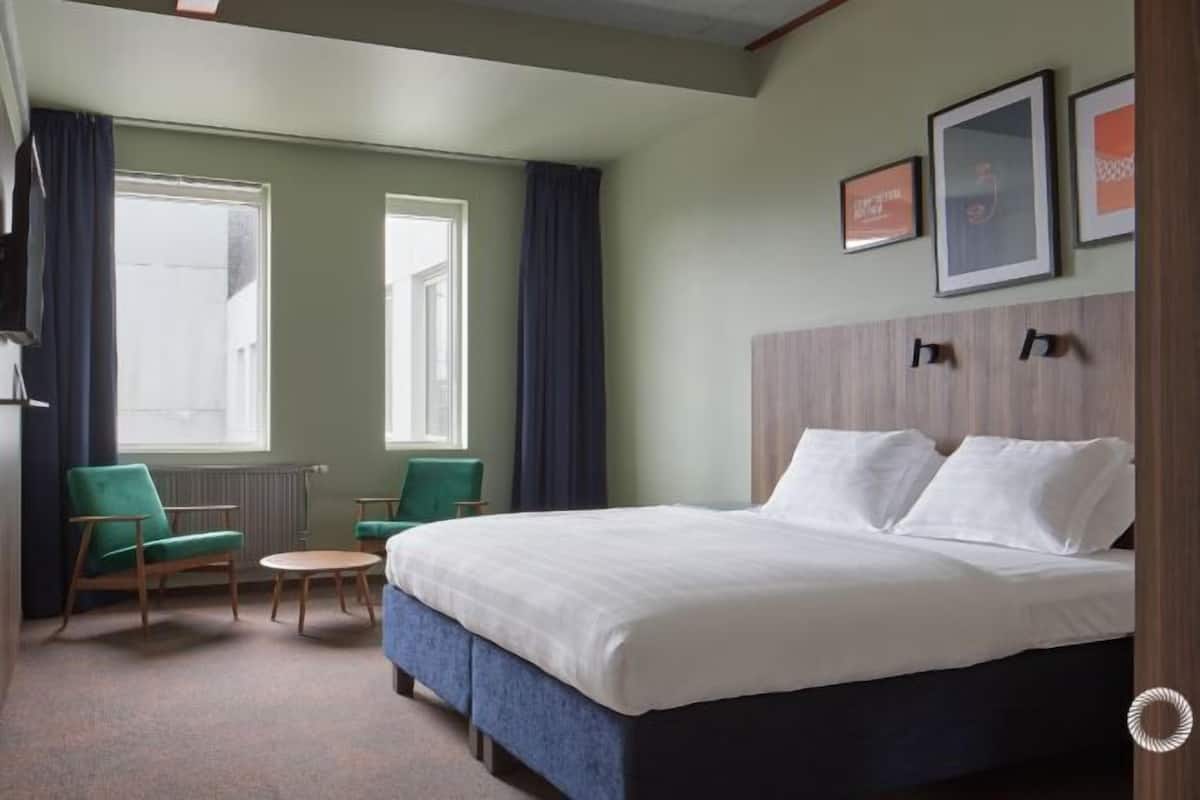 Ein Hotelzimmer mit Bett, zwei Stühlen, einem kleinen Tisch und einem Fenster mit Vorhängen.