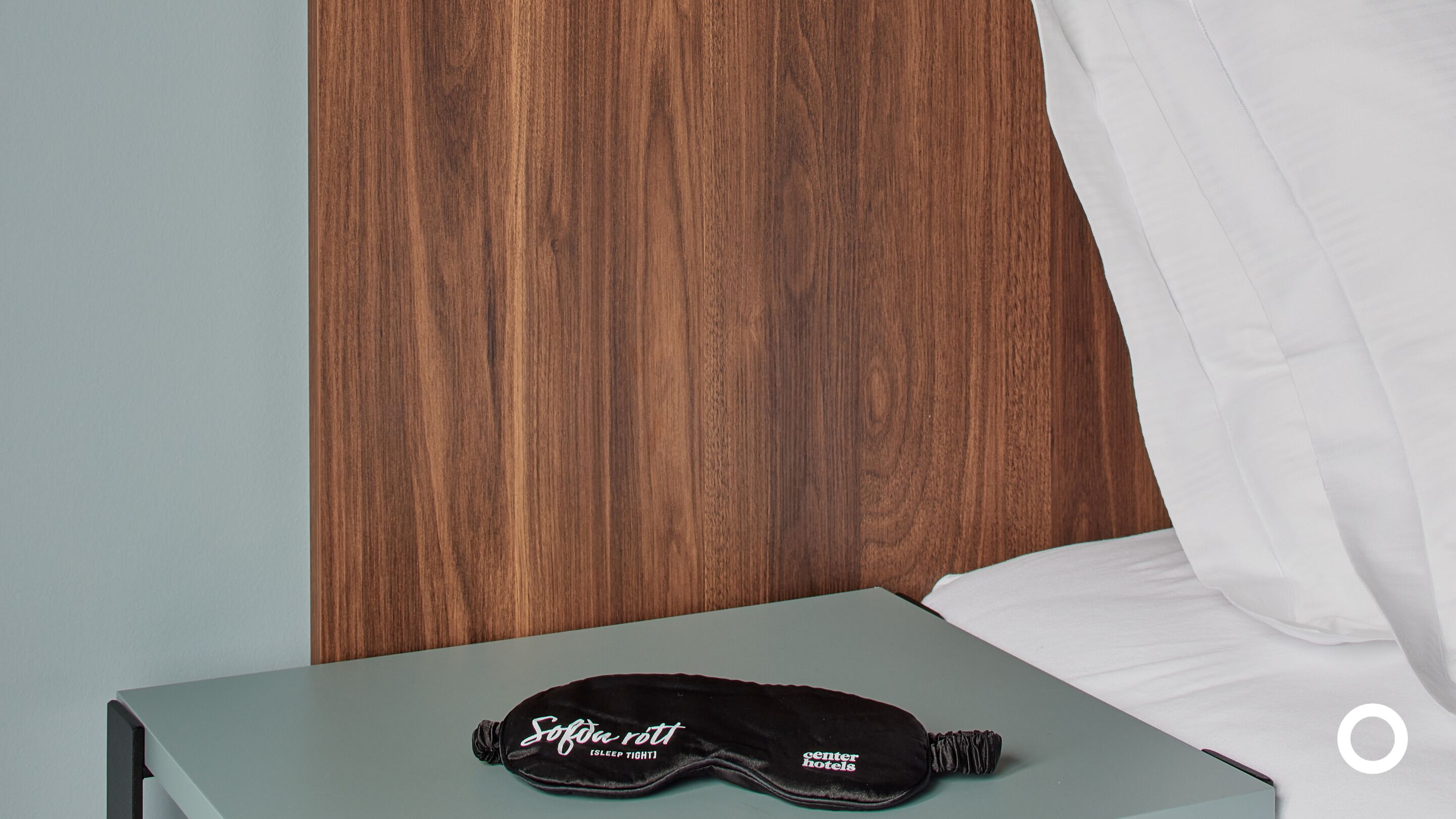 A bed with a wooden headboard, a pillow, and a sleep mask on a bedside table.