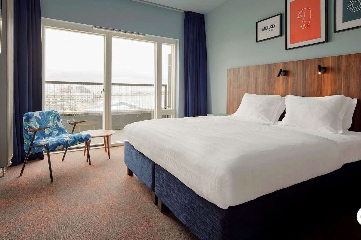 Ein Hotelzimmer mit einem großen Bett, einem Sessel mit tropischem Muster, einem kleinen Tisch und Blick auf die Stadt durch große Fenster.