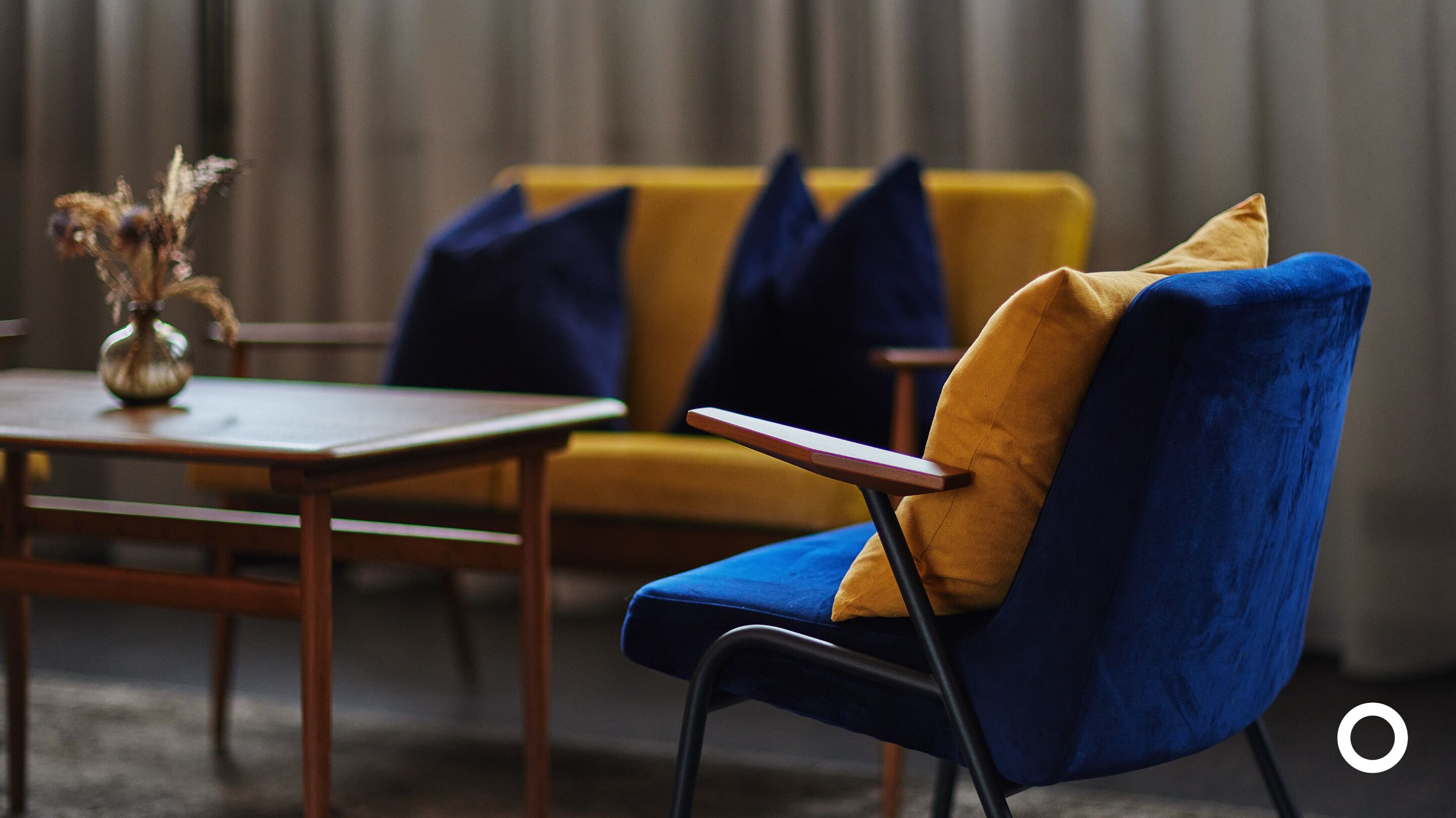 A modern living room with a blue and yellow sofa, a wooden coffee table, and a vase with dried flowers.