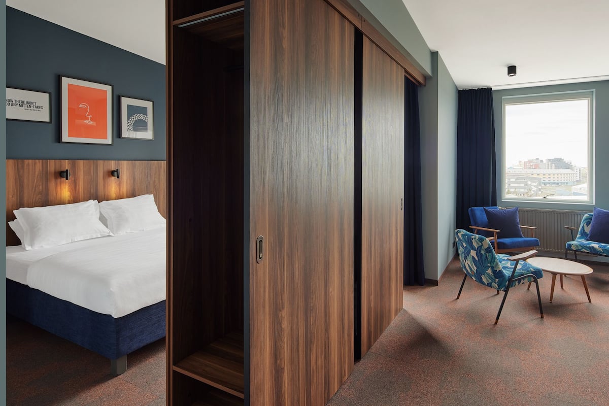 Ein Hotelzimmer mit Bett, einem Schrank, einer Sitzecke mit zwei Sesseln und Blick auf die Stadt durch große Fenster.
