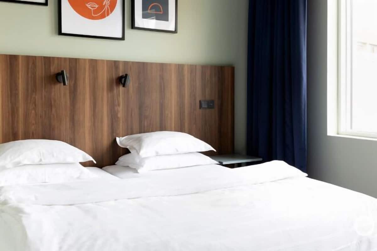 Ein Hotelzimmer mit einem hölzernen Kopfteil, zwei Betten mit weißer Bettwäsche und Kissen, einem Nachttisch und einem Fenster mit Vorhängen.