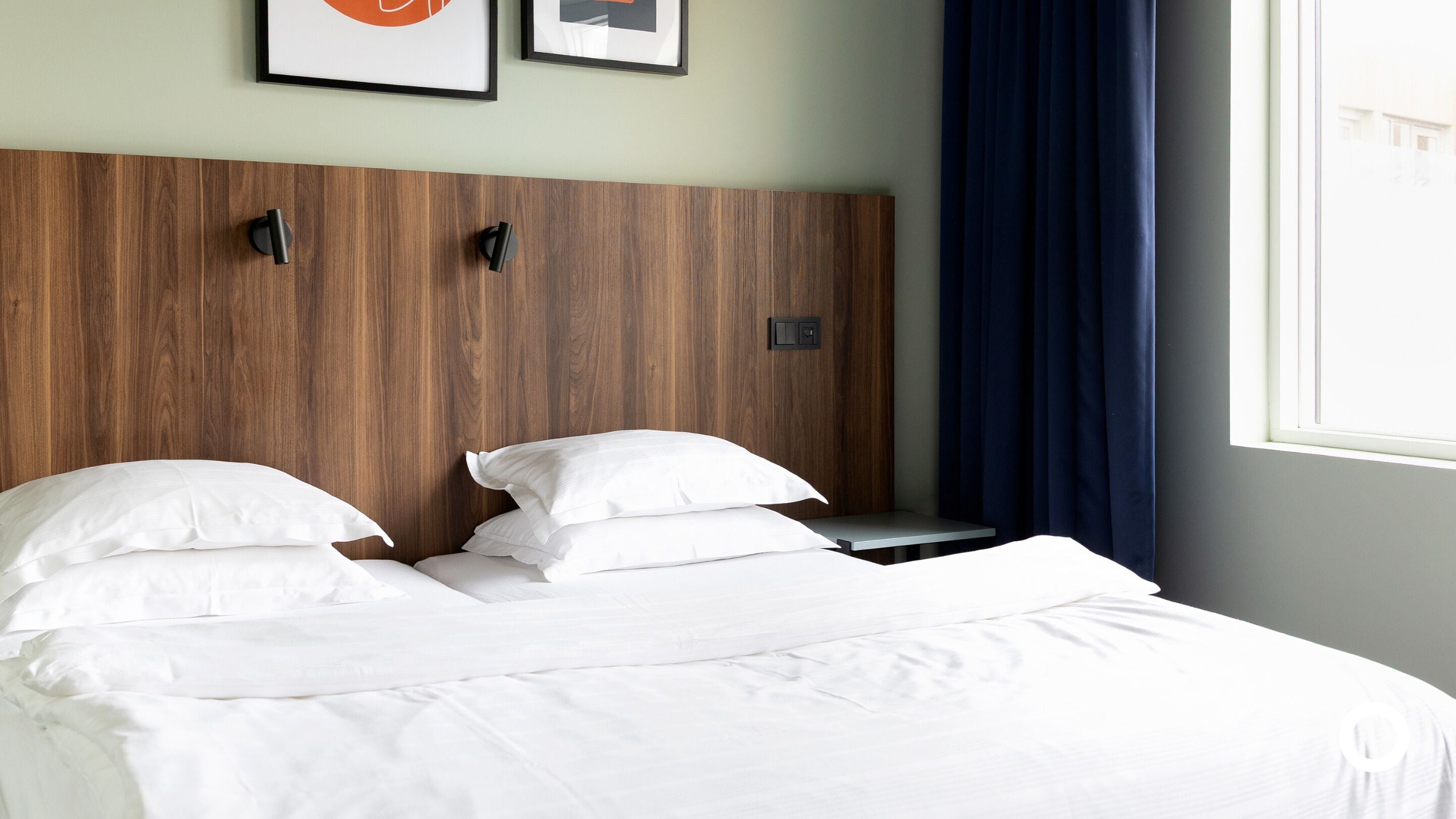 A neatly made bed with white linens and pillows, a wooden headboard, a bedside table, and a window with blue curtains.