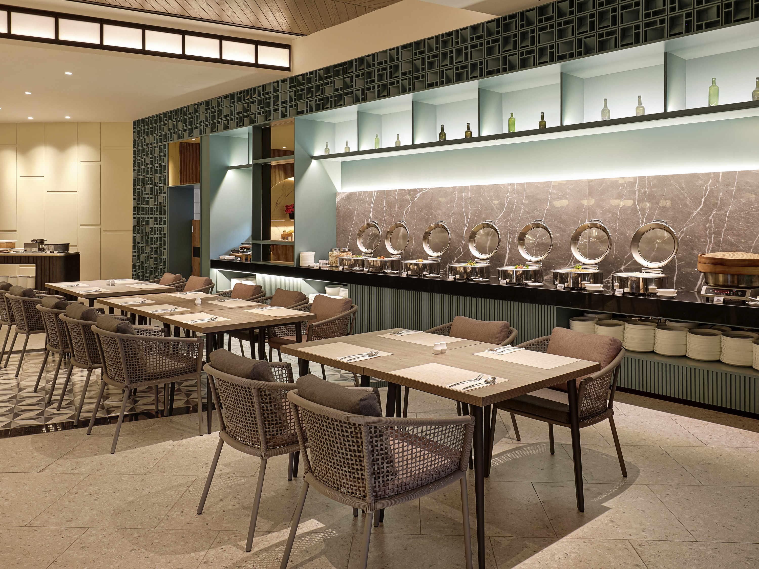 A modern restaurant with multiple dining tables and chairs, a buffet counter with various food items, and a decorative wall with a geometric pattern.