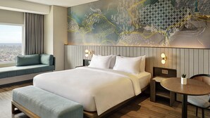 Una habitación de hotel moderna con una cama grande, un área de descanso junto a la ventana, una mesa redonda y un mural decorativo en la pared.