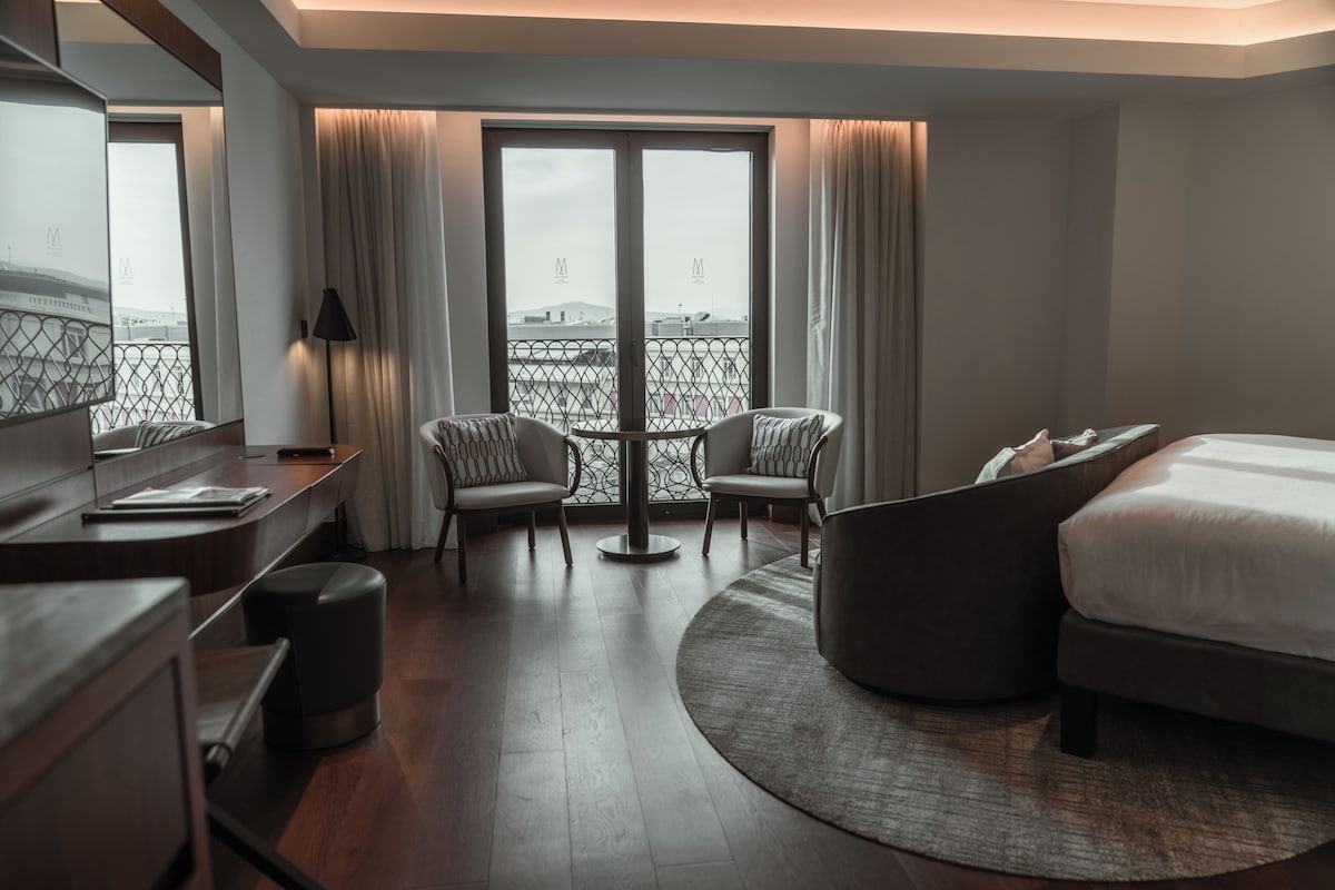 A hotel room with a bed, two armchairs, a desk, and a large window with a view.