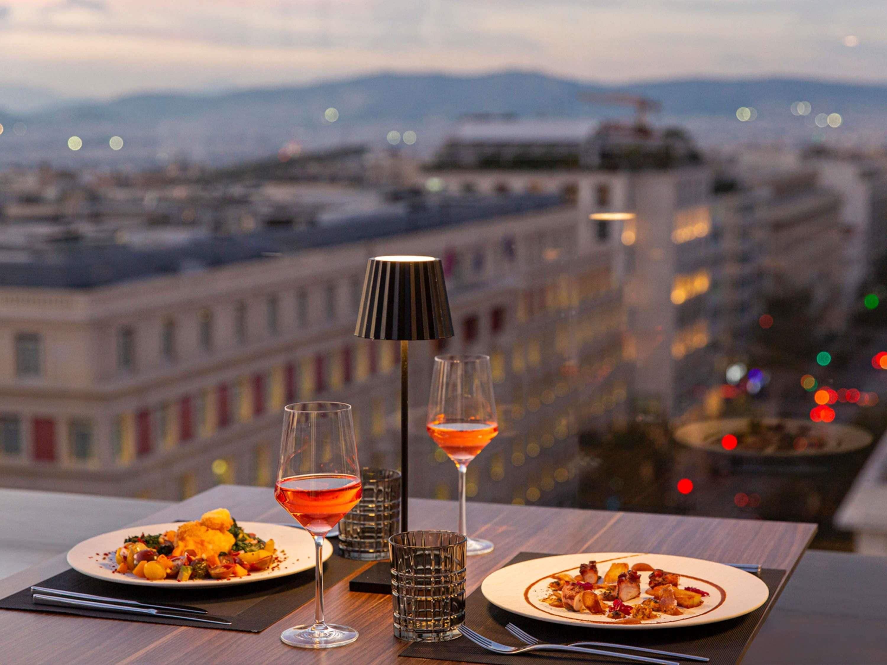 Uma mesa com dois pratos de comida, dois copos de vinho e um abajur em um terraço com vista para a paisagem urbana.