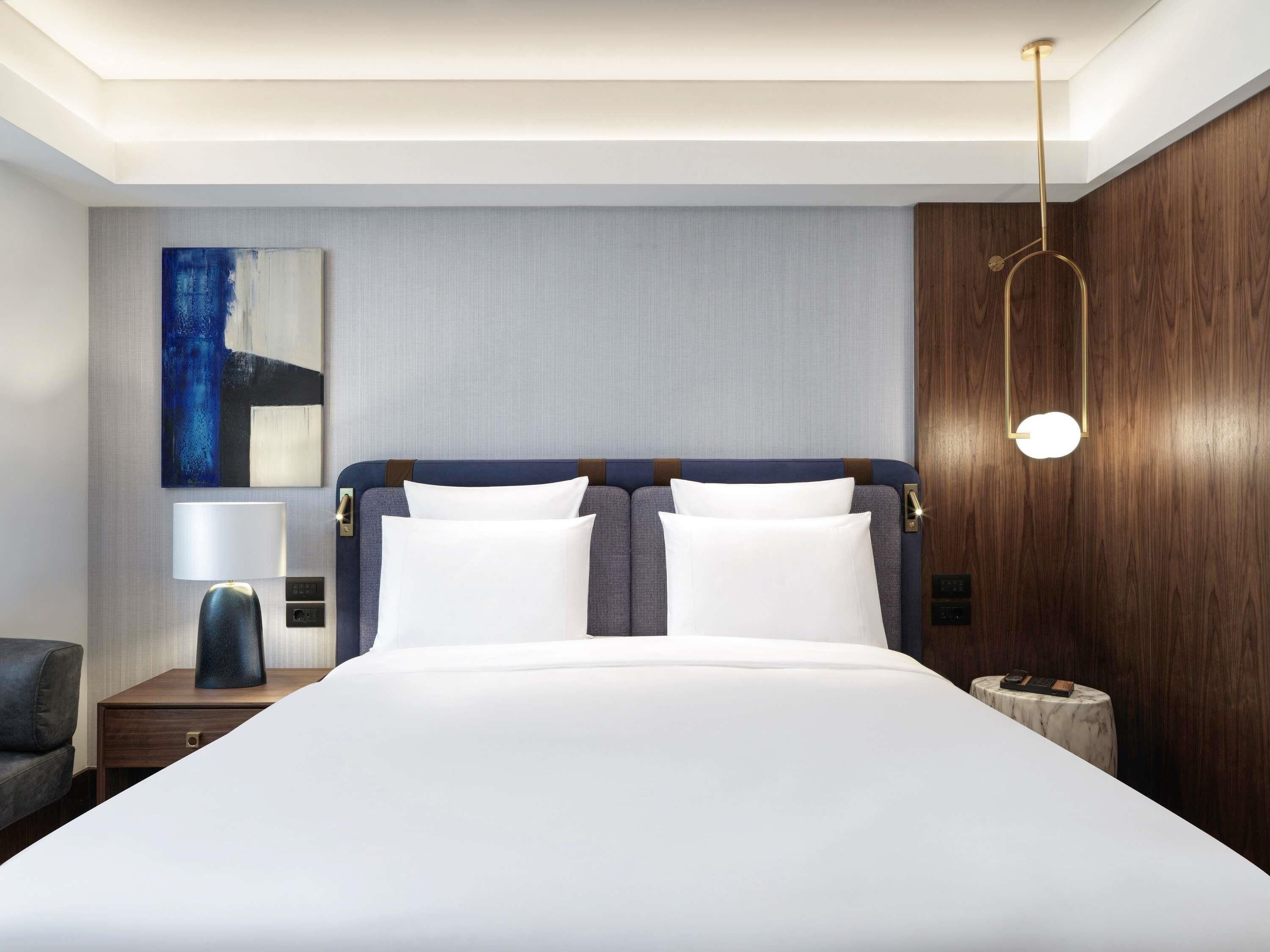 Ein modernes Hotelzimmer mit einem großen Bett, einem Nachttisch mit Lampe, einer Wand mit Holzvertäfelung und einer Deckenleuchte.