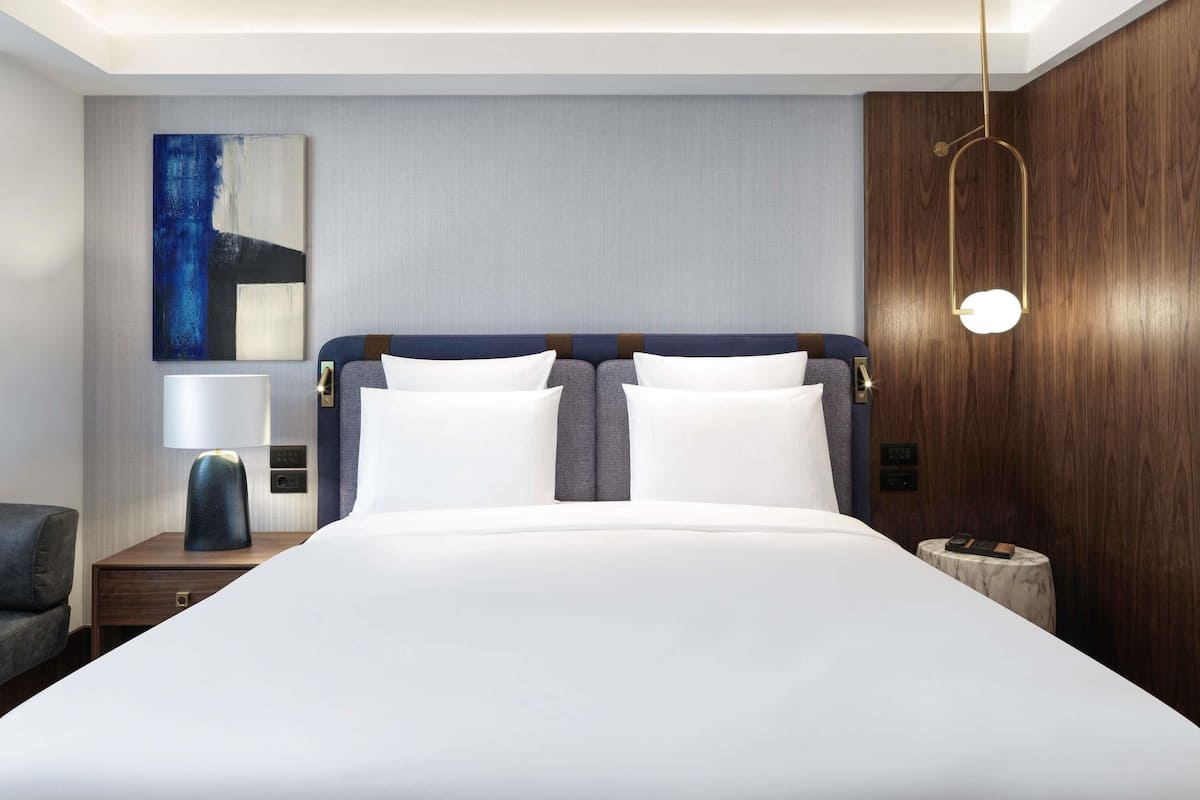 Ein modernes Hotelzimmer mit einem großen Bett, einem Nachttisch mit Lampe, einer Wand mit Holzvertäfelung und einer Deckenleuchte.