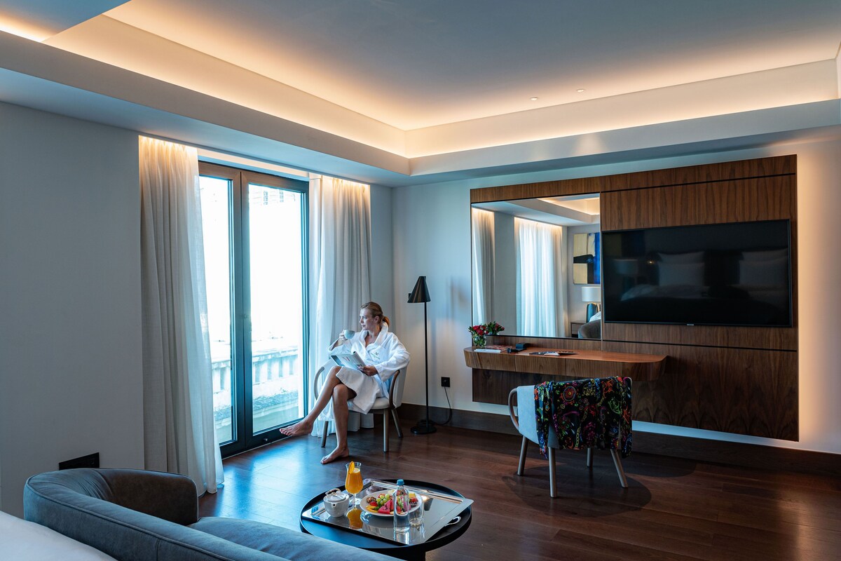 A modern hotel room with a large bed, a sitting area with a sofa, a dining table with breakfast, and a TV mounted on the wall.