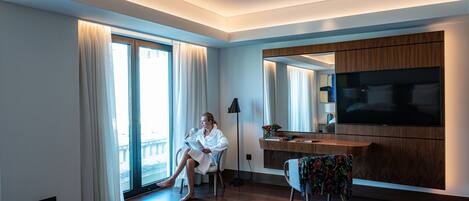 A modern hotel room with a large bed, a sitting area with a sofa, a dining table with breakfast, and a TV mounted on the wall.