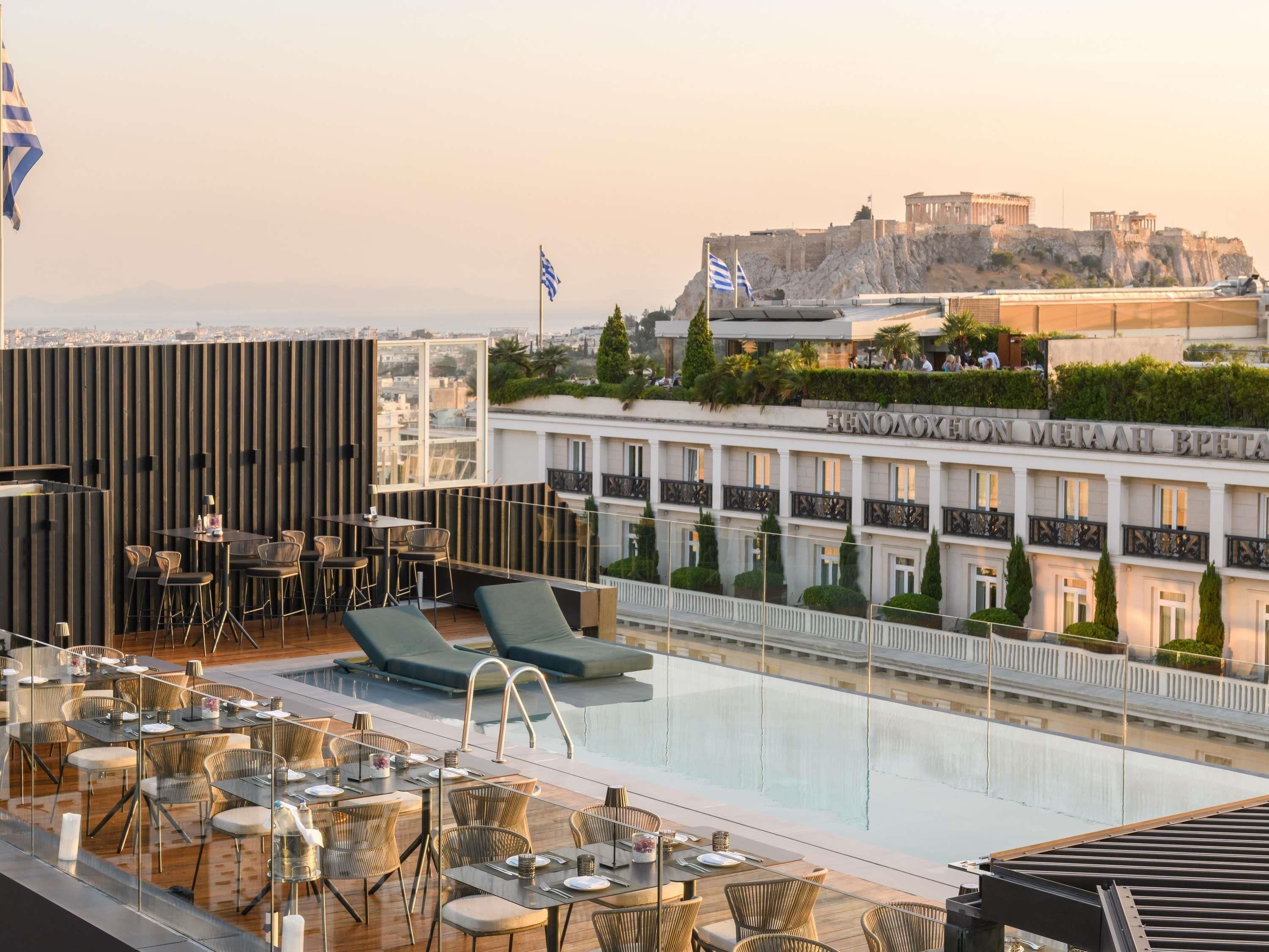 Área de piscina no terraço, com espreguiçadeiras e mesas, com vista para um edifício histórico com bandeiras.
