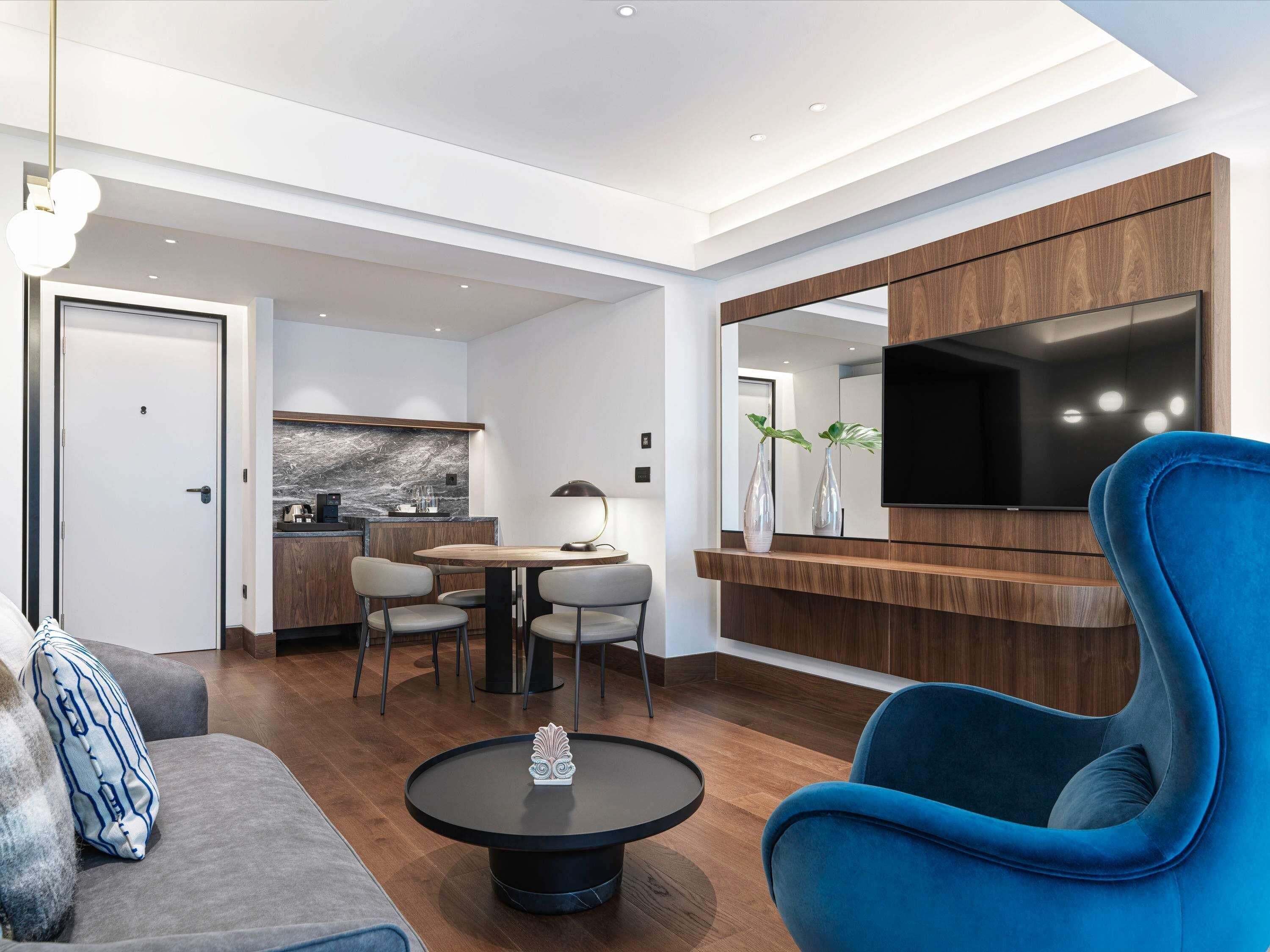 Una sala moderna con un sofá, un sillón azul, una mesa de centro, una zona de comedor con sillas, una cocina al fondo y un televisor montado en un panel de madera.