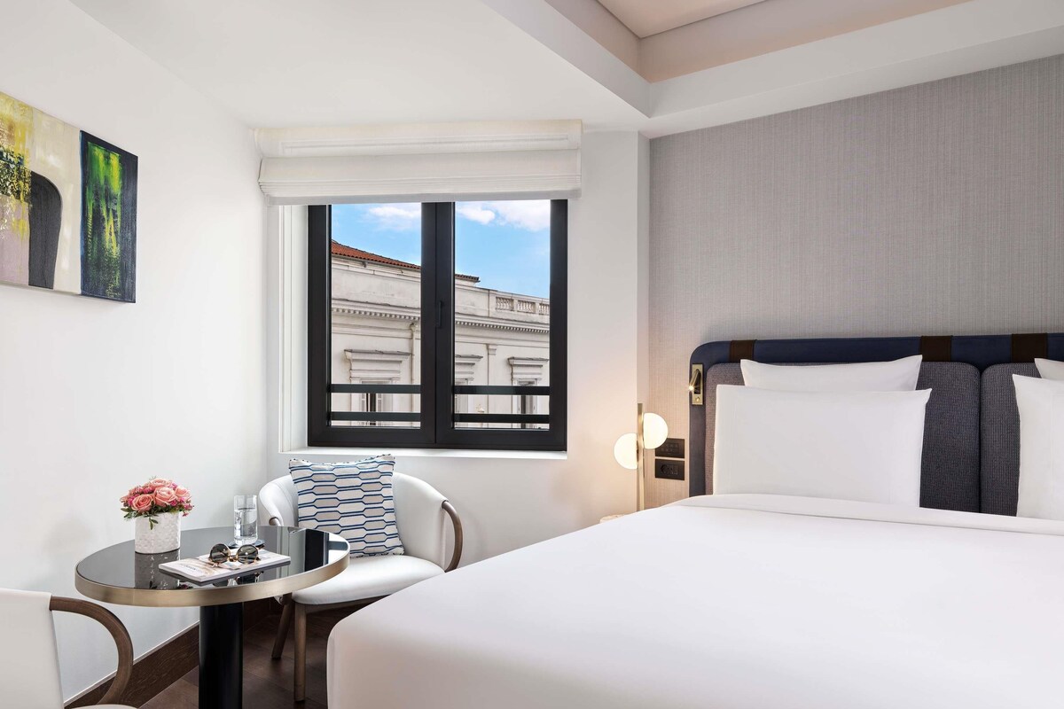 Habitación de hotel con cama, una mesa redonda con una silla, una ventana con vistas a edificios y una pared decorada con obras de arte.