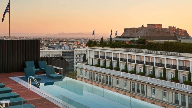 A rooftop pool with lounge chairs, a view of a historic building, and flags. - Athens Capital Center Hotel-MGallery Collection (Athens)