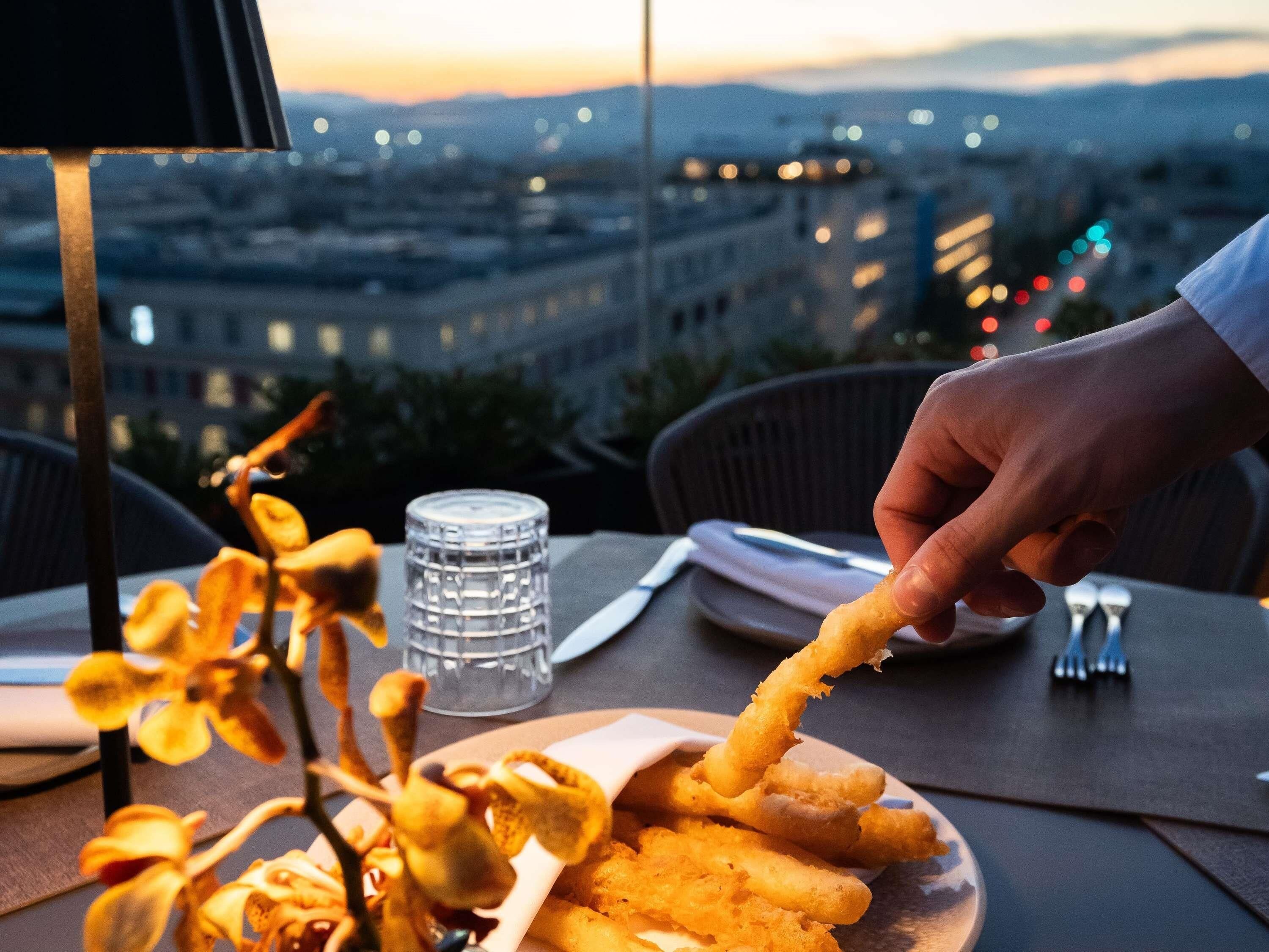 Una persona sosteniendo un alimento, con un plato de elementos similares y un vaso de agua sobre una mesa. Al fondo, se observa una vista de la ciudad al anochecer.