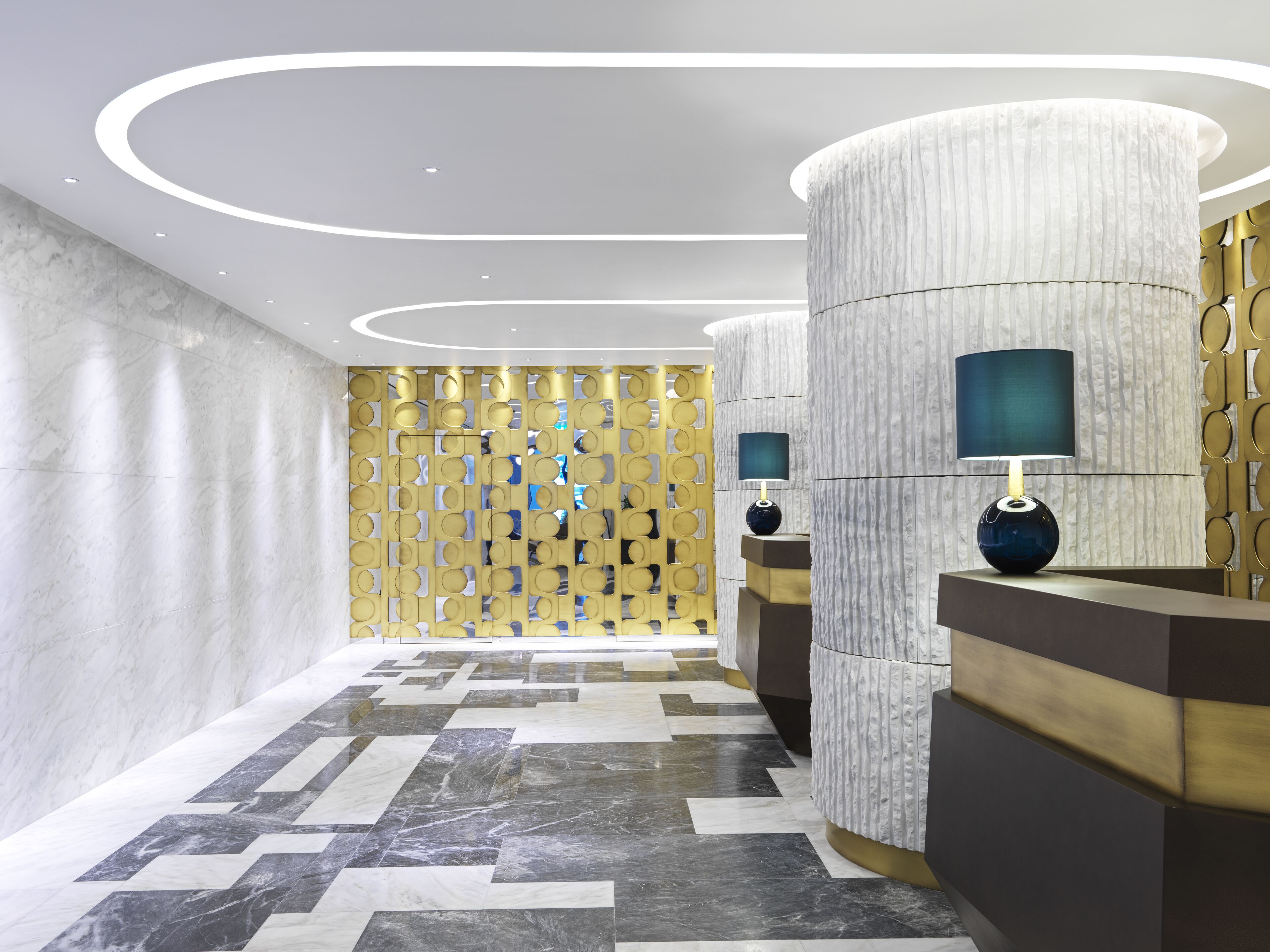 A modern hotel lobby with marble flooring, a patterned wall, and two cylindrical columns with lamps.