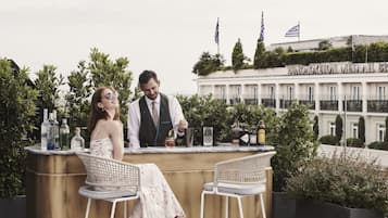 Um bar no terraço com duas cadeiras de vime branca, um bartender e um balcão com diversos copos e garrafas.
